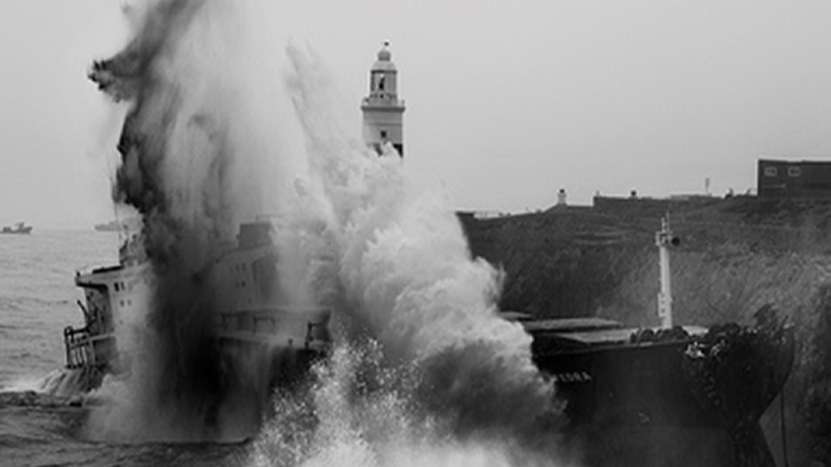 A dramatic monochrome photograph captures MV Fedra being battered by waves as it smashes against the rocky cliffs of Europa Point, Gibraltar. The lighthouse in the background highlights the ship’s proximity to land during the grounding incident. This image illustrates the extreme weather conditions that contributed to the vessel's structural failure and subsequent oil spill.