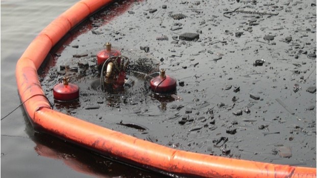 An oil spill containment boom surrounding heavily oiled water with a disc skimmer in operation, demonstrating in-situ oil recovery efforts at Karachi Port.