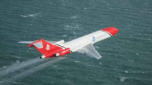 Aerial view of OSRL's Boeing 727 in flight, actively releasing dispersant over open water. Emphasises operational readiness and real-world deployment capability.