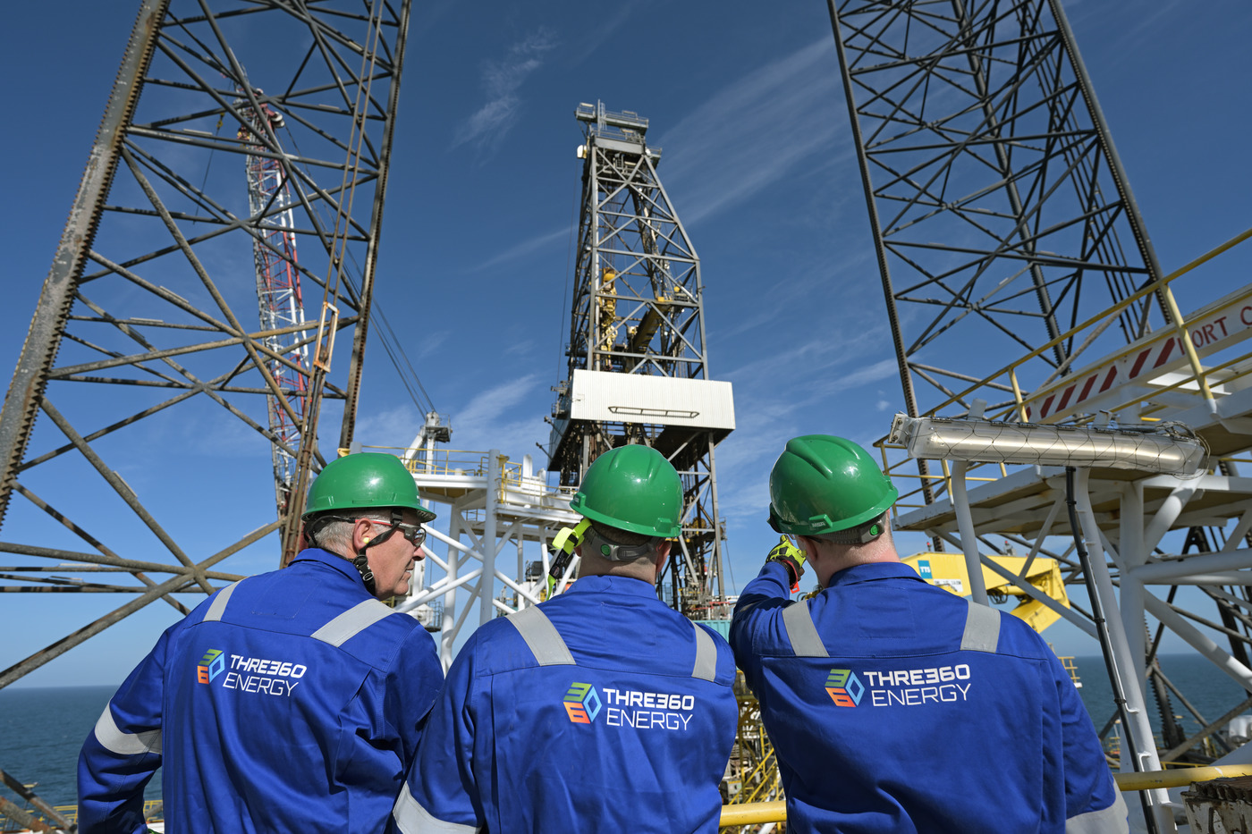 Three THREE60 Energy engineers in green helmets assess offshore rig infrastructure beneath a clear sky. The image captures teamwork, engineering, and scale.