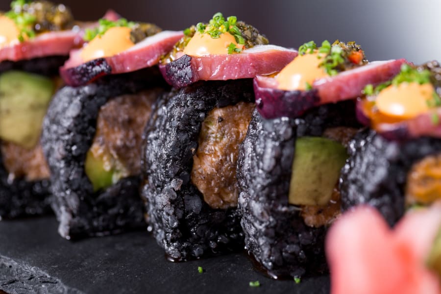 Black rice sushi rolls topped with seared tuna and sauce, shot close on slate.
