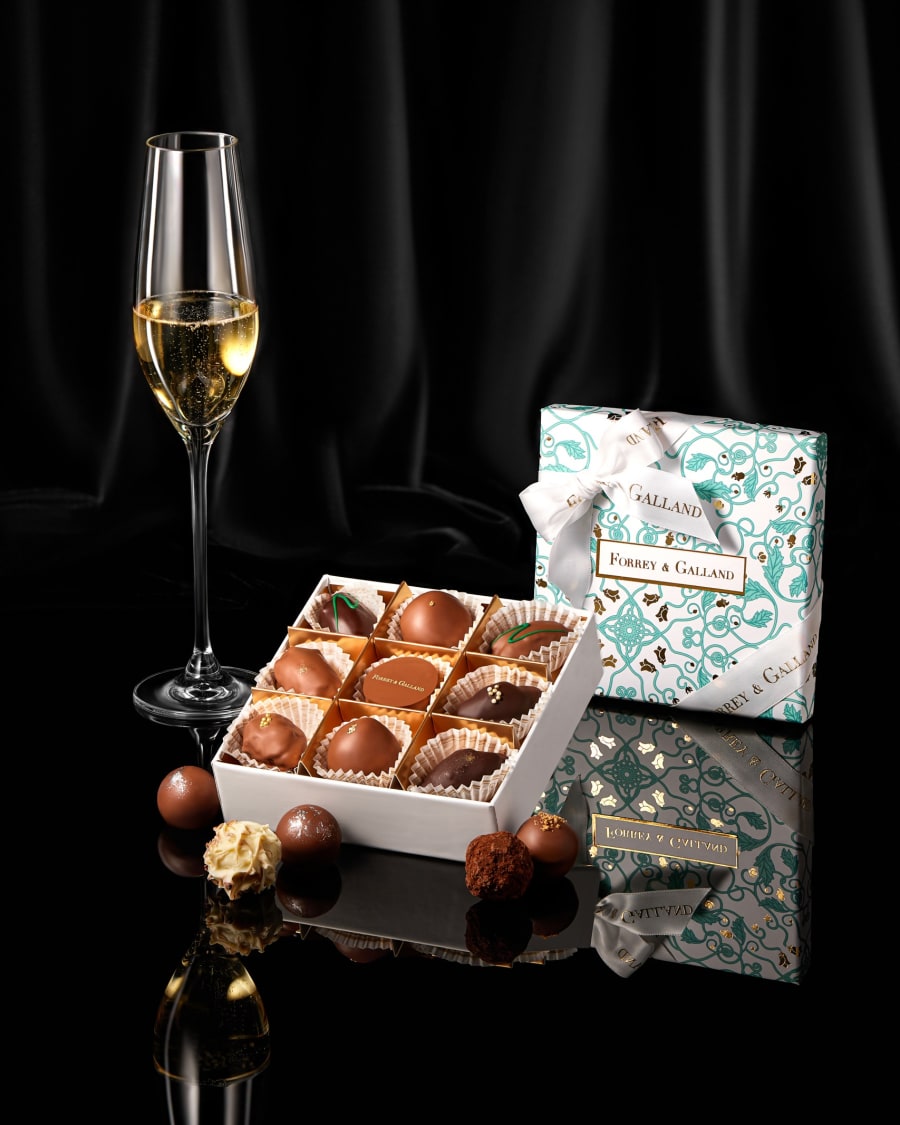 A box of handmade chocolates with a glass of sparkling drink on black velvet.