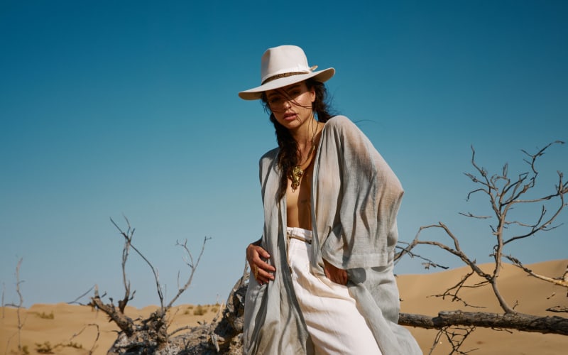 A model in a hat and light layered outfit posing in the desert under a blue sky.