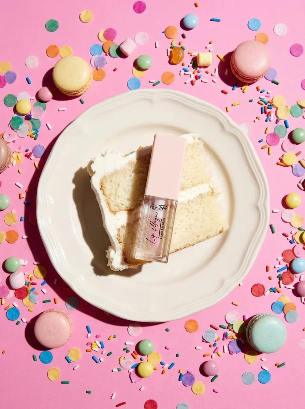 A lip gloss standing on a slice of cake among macarons and confetti — a packshot extended into an editorial scene.