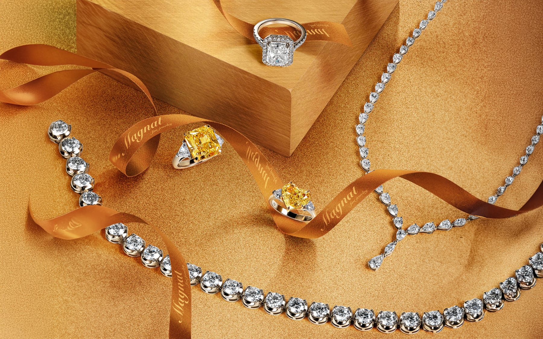 Diamond rings and necklaces staged on gold sand with a branded satin ribbon for a jewelry campaign.
