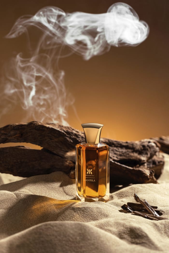 A perfume bottle on sand with smoke drifting across the frame.