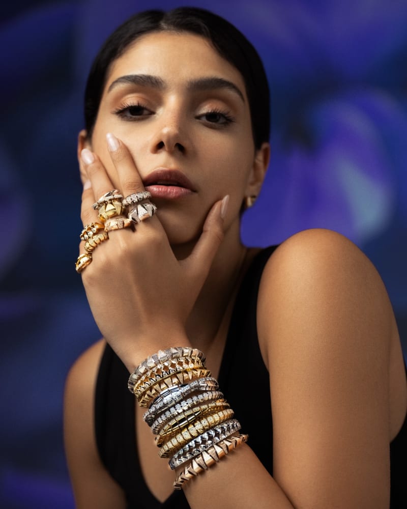 Stacked gold rings and bangles worn on a model's hands against a blue backdrop.