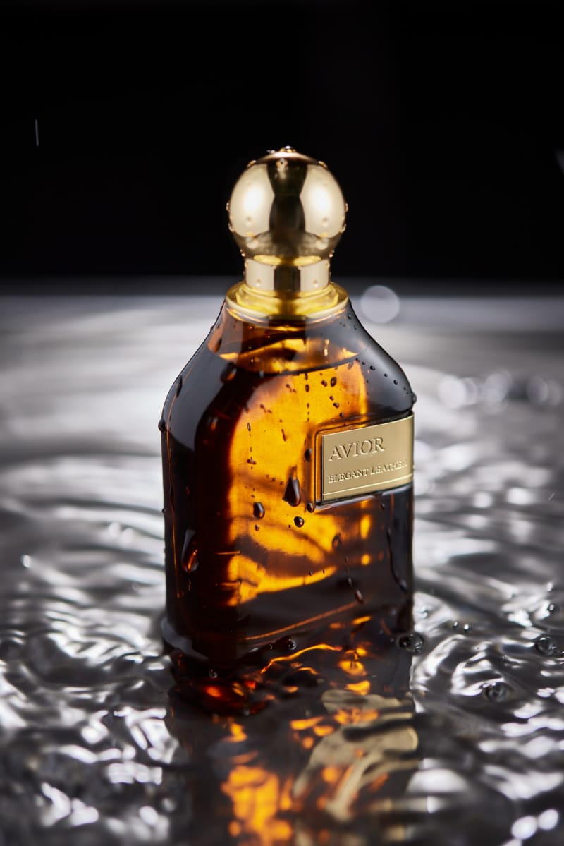 A dark amber perfume bottle with a gold cap standing in rippling water, droplets suspended around it.