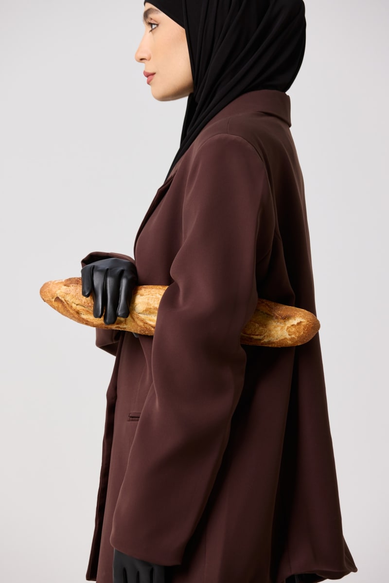 A model in a hijab and brown blazer holding a baguette under a leather-gloved arm.