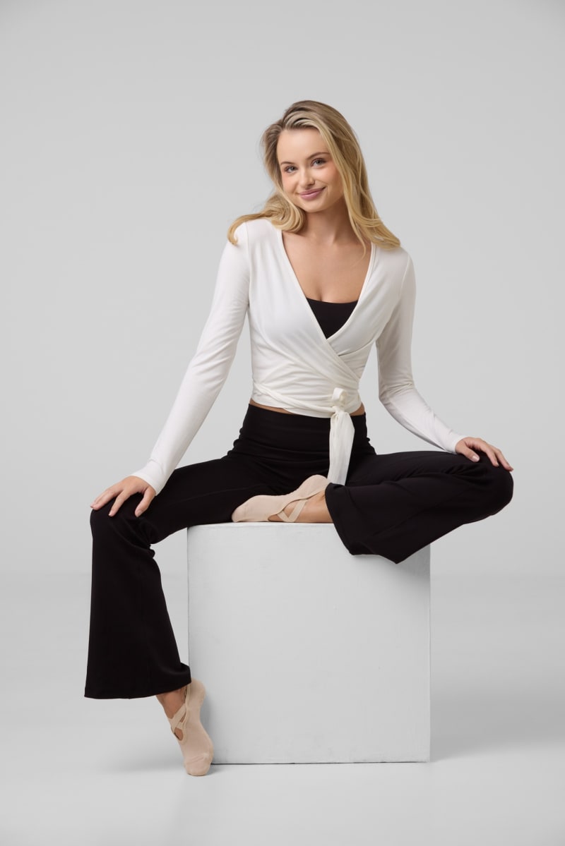 A model in a white wrap top and black trousers seated on a white cube.