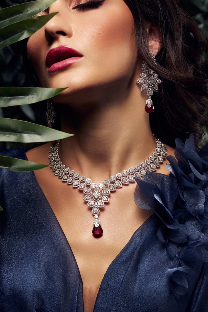 A diamond and ruby necklace with matching earrings, worn with a navy gown among palm leaves.