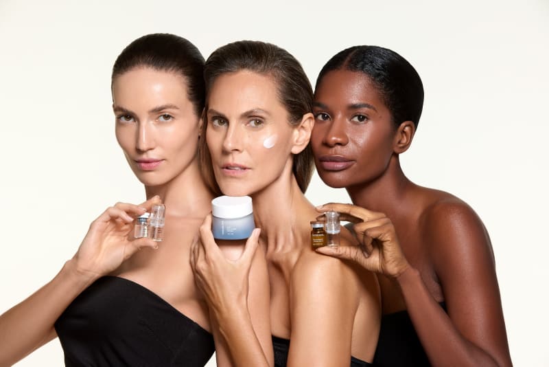 Three models of different skin tones holding skincare vials and a jar, one with cream on her cheek.