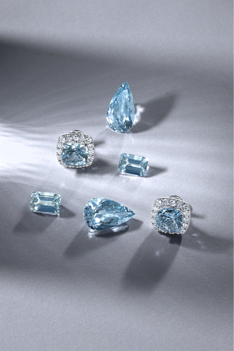Loose aquamarine stones and halo studs arranged on grey under hard light with long shadows.