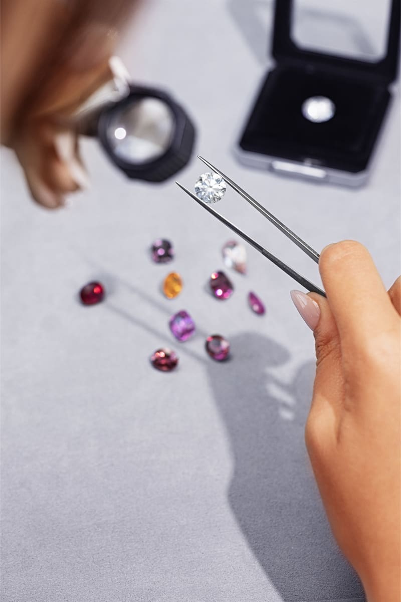 A gemologist's hands holding a loupe and tweezers with a round brilliant diamond, loose coloured stones on the bench.