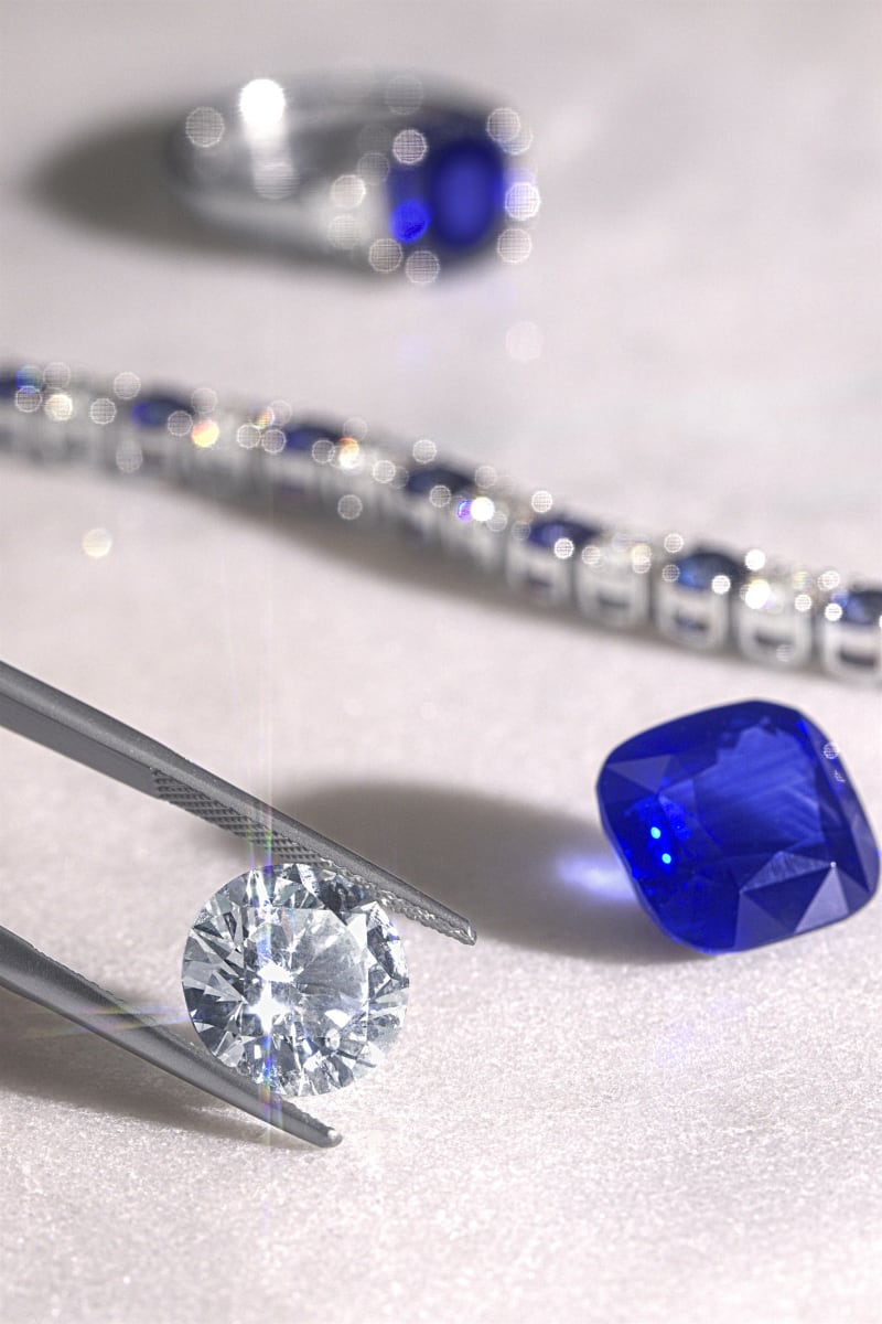 Tweezers holding a round brilliant diamond in macro, a blue sapphire and a sapphire tennis bracelet behind.