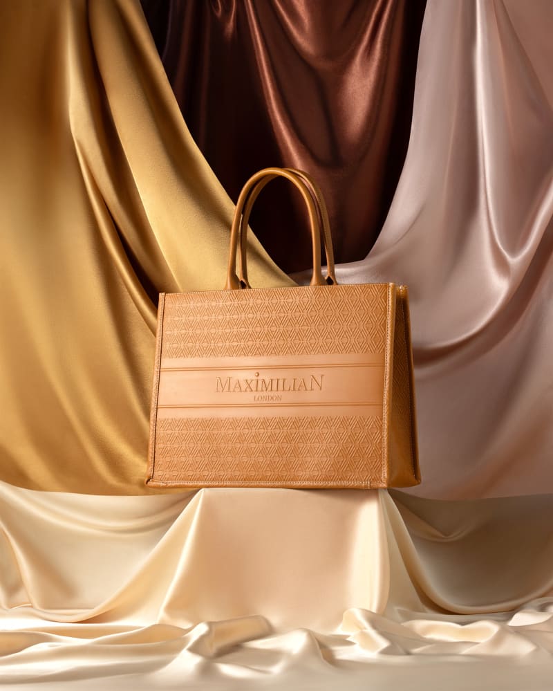 A tan leather tote standing on cream silk against draped gold and brown fabric.