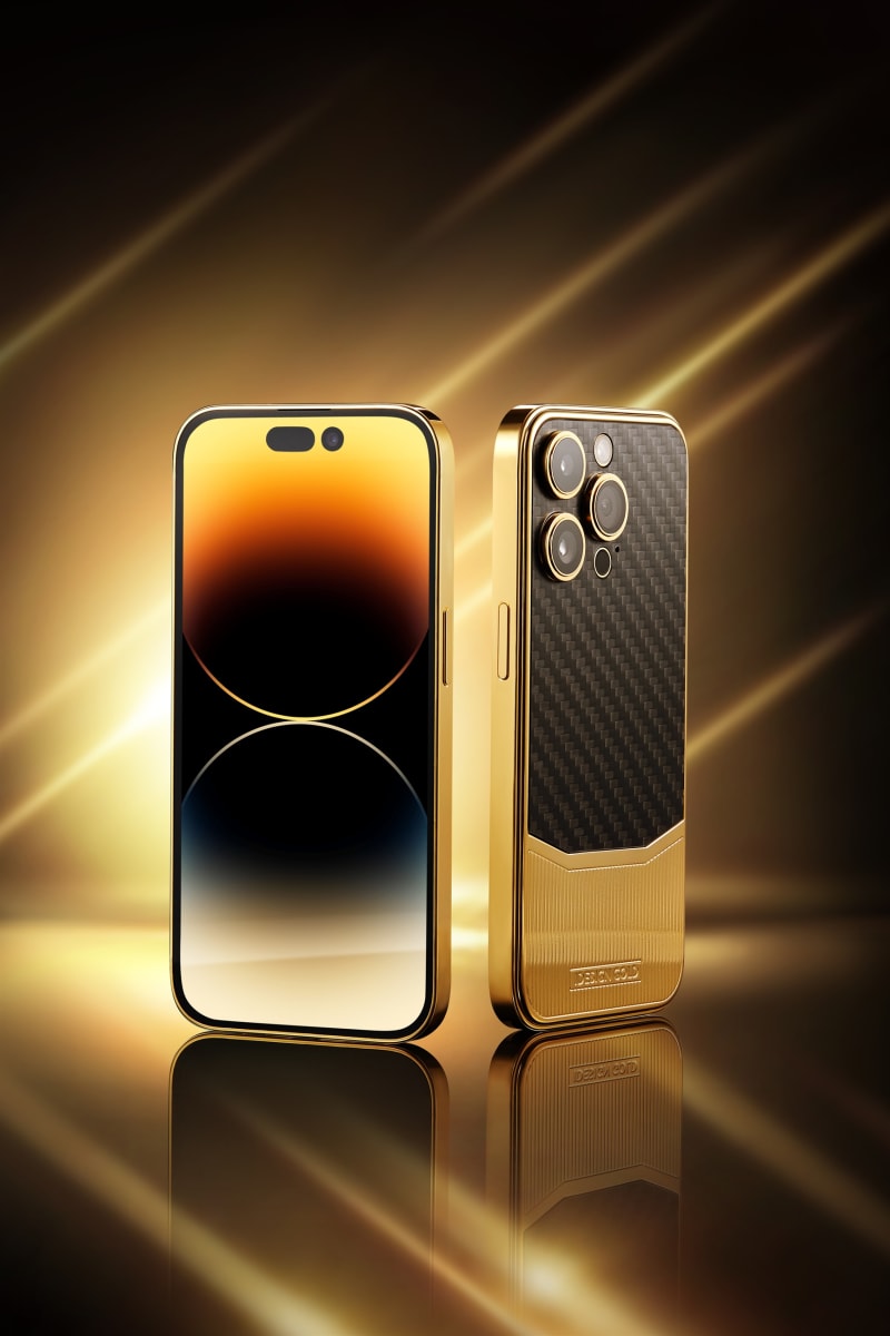 A gold iPhone and a carbon-fibre case standing on a mirrored surface under streaks of golden light.