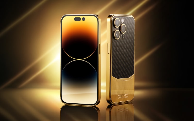 A gold iPhone shown front and back with a carbon-fibre case, on a mirrored surface under streaks of golden light.