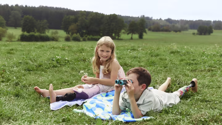 A boy with a prosthesis and binoculars is lying on the grass next to a girl with a prosthesis who is sitting down.