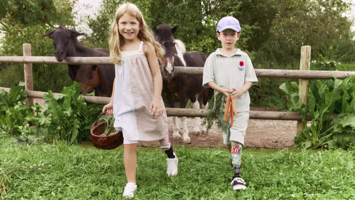 A girl with a prosthesis and a boy with a prosthesis walk side by side, with a horse in the background.