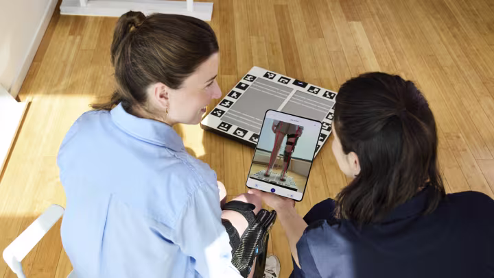A technician showing a patient the alignment results on a tablet. The Ottobock lazar posture measuring plate sits on the floor in the background. 