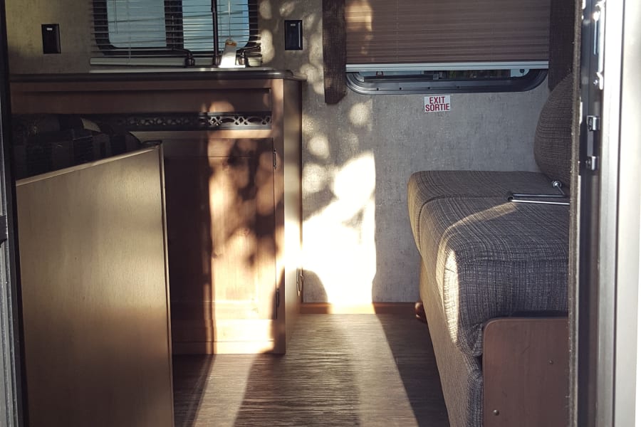 An RV rental interior shows a kitchen, living area, and sunlit wood floors with an "Outdoors RV" mat.