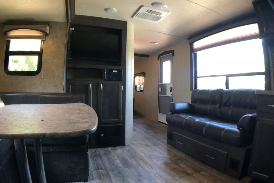 Spacious RV rental interior with a dining table & booth, black leather sofa, entertainment center, and wood-look flooring.