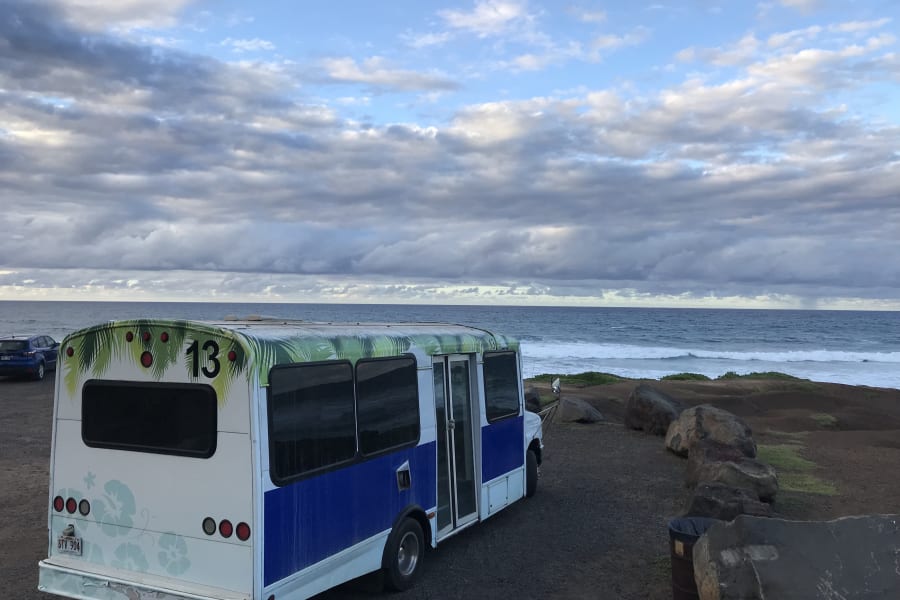 Hawaii RV rental bus (13) by ocean waves under a cloudy sky. Scenic vehicle for island travel.