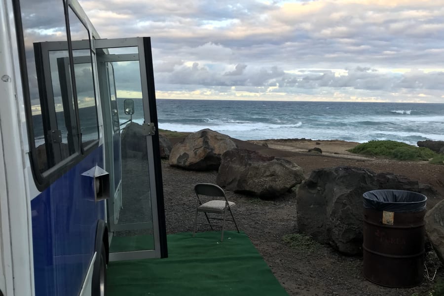 RV rental bus parked on a gravel coast, door open with green mat & chair, offering ocean views under a cloudy sky.