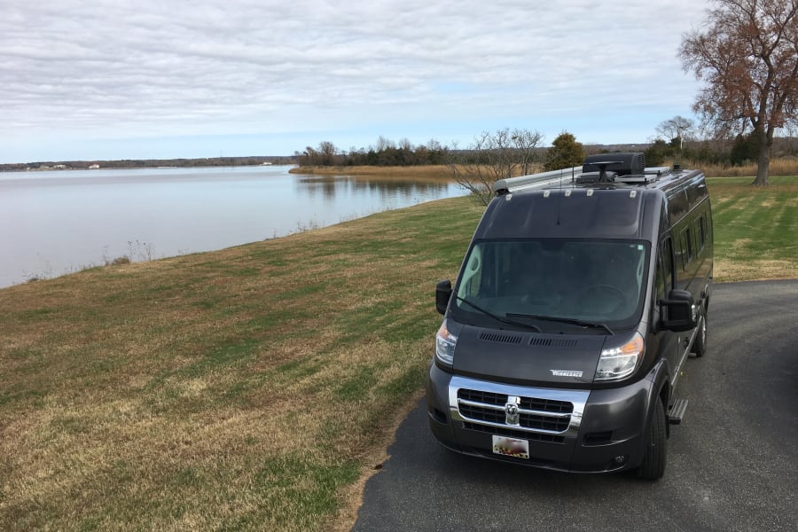 A grey Winnebago RV van is parked on asphalt beside a grassy slope leading to a calm bay under a cloudy sky, ideal for an RV rental.