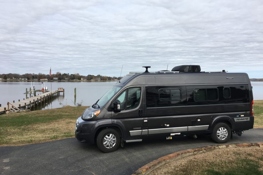 This dark gray camper van, a potential RV rental, is parked beside a wooden pier extending into a calm bay. Overcast sky.