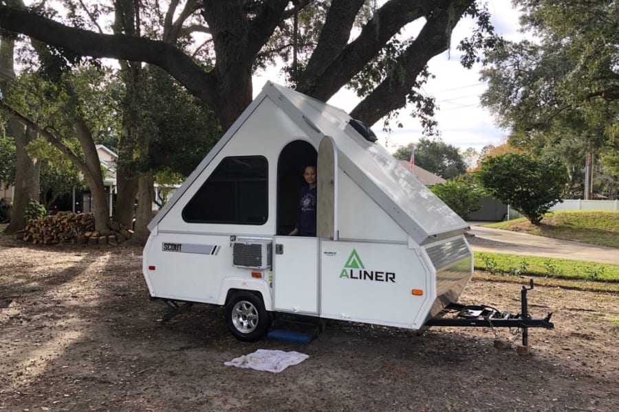A person smiles from inside a white Aliner A-frame RV rental, parked on a leafy ground under large trees.