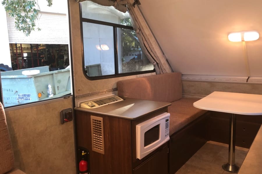 Inside an RV rental: a compact living space featuring a dining booth with cushions, table, microwave, and exterior view.