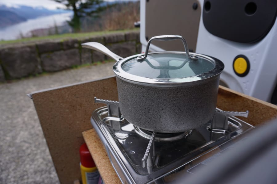 A speckled gray pot with a glass lid sits on a portable gas stove, part of an RV rental kitchen setup with a scenic outdoor view.