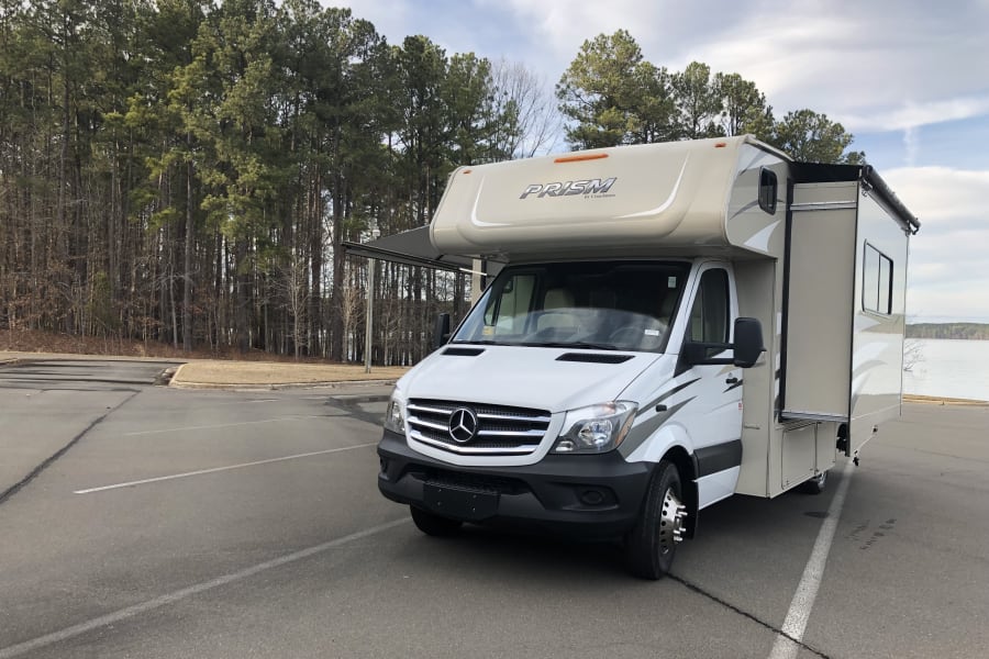 A white and beige Coachmen Prism Class C RV rental with an extended slide and awning, parked by a forest and lake.