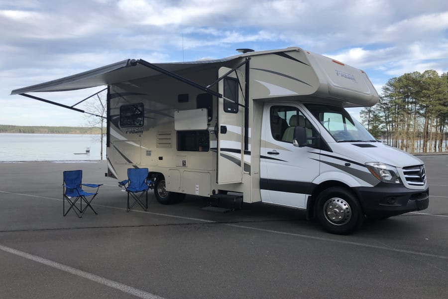 This RV rental, a Class C motorhome, is lakeside with an extended awning & blue chairs for relaxation.