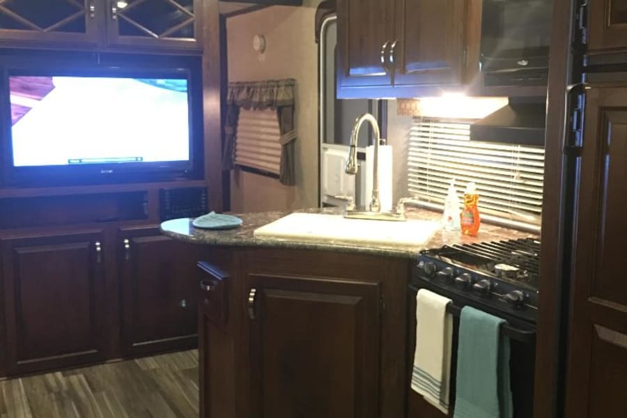 Modern rv rental interior: full kitchen with dark wood cabinets, sink, stovetop, microwave, and cozy TV living area.