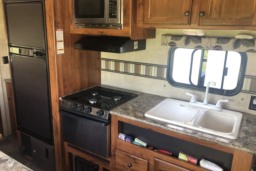 This RV rental kitchen features a refrigerator, microwave, stove, oven, and double sink with wooden cabinets.