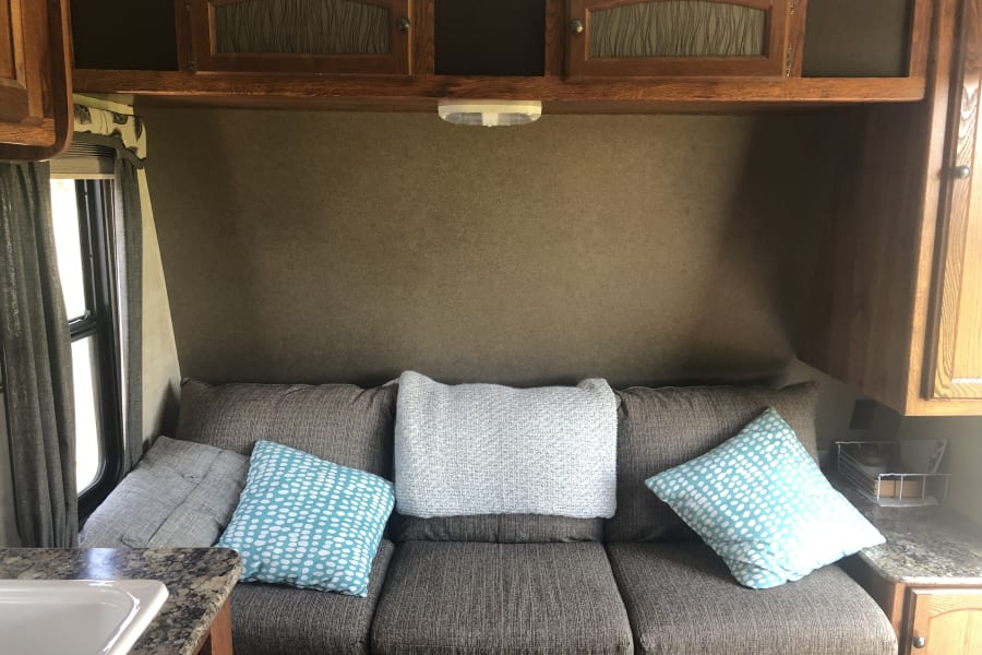 Comfortable brown couch with blue and white patterned pillows inside an RV rental, featuring wood cabinets and a window.