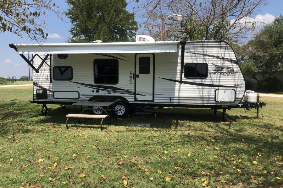 This Jay Flight travel trailer, an RV rental, is parked at a sunny campsite with a picnic table and autumn leaves on the grass.