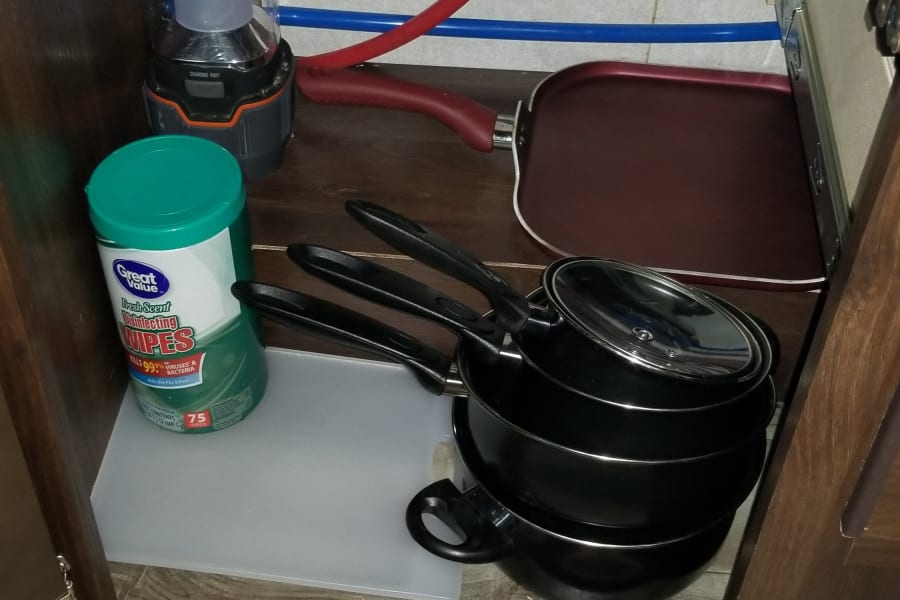 RV rental kitchen cabinet with stacked pots, griddle, wipes, portable light, and cutting board.