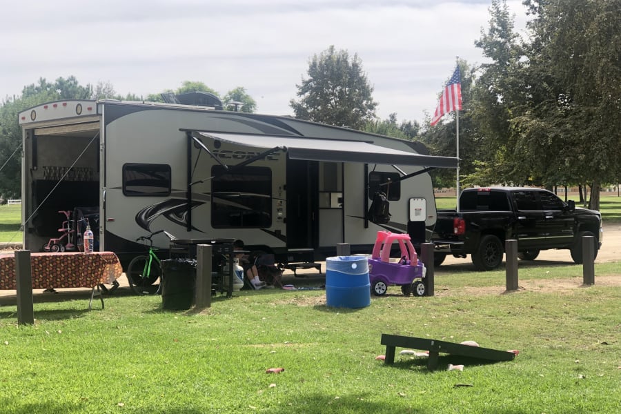 RV rental campsite with gray RV, extended awning, truck, picnic table, toys, cornhole, and an American flag.