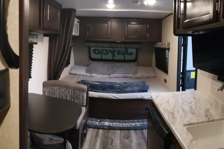 Inside an RV rental: view of a bed, dinette, kitchen counter with sink, and overhead cabinets.