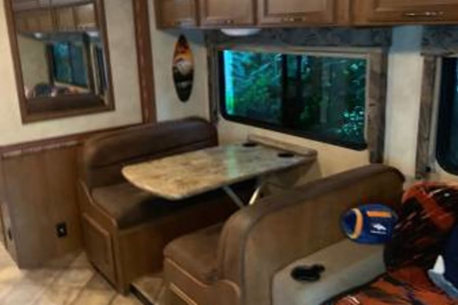 Inside this RV rental: a tiled floor leads to a brown dining booth, table, and storage cabinets, with a comfy couch nearby.