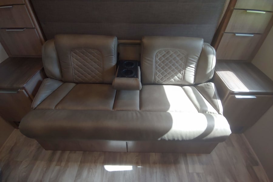 Comfortable RV rental interior featuring a tan dual recliner sofa with cup holders and adjacent storage cabinets.