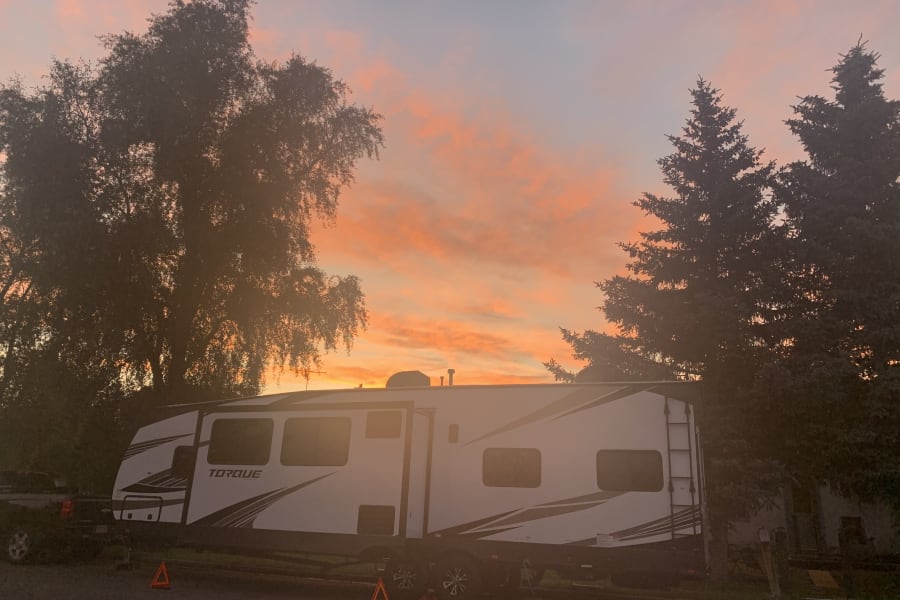 Vibrant orange and pink sunset sky above an RV rental, parked with a truck between silhouetted trees.