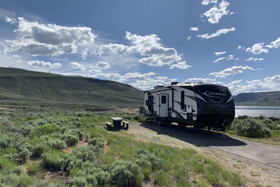 RV rental parked at a scenic lakeside campsite, with green hills, a picnic table, and blue sky.