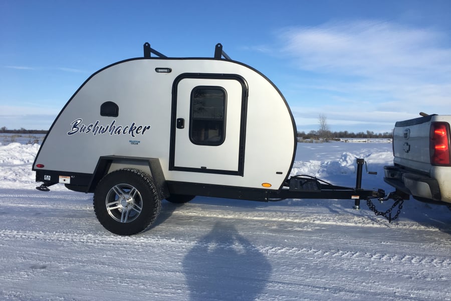 White Bushwhacker teardrop RV rental hitched to a truck on a snowy landscape under a clear blue sky.