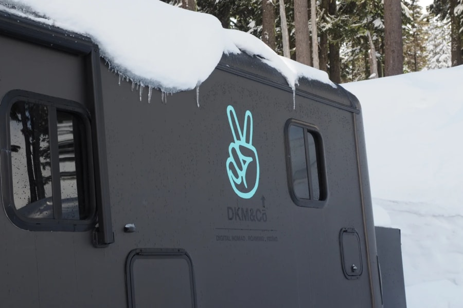Dark gray RV rental in a snowy forest, with ice and a peace sign logo. Ideal for digital nomads.