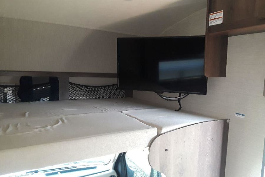 Inside an rv rental: a cozy over-cab bunk bed with a mounted TV and storage nets, ideal for travel comfort.